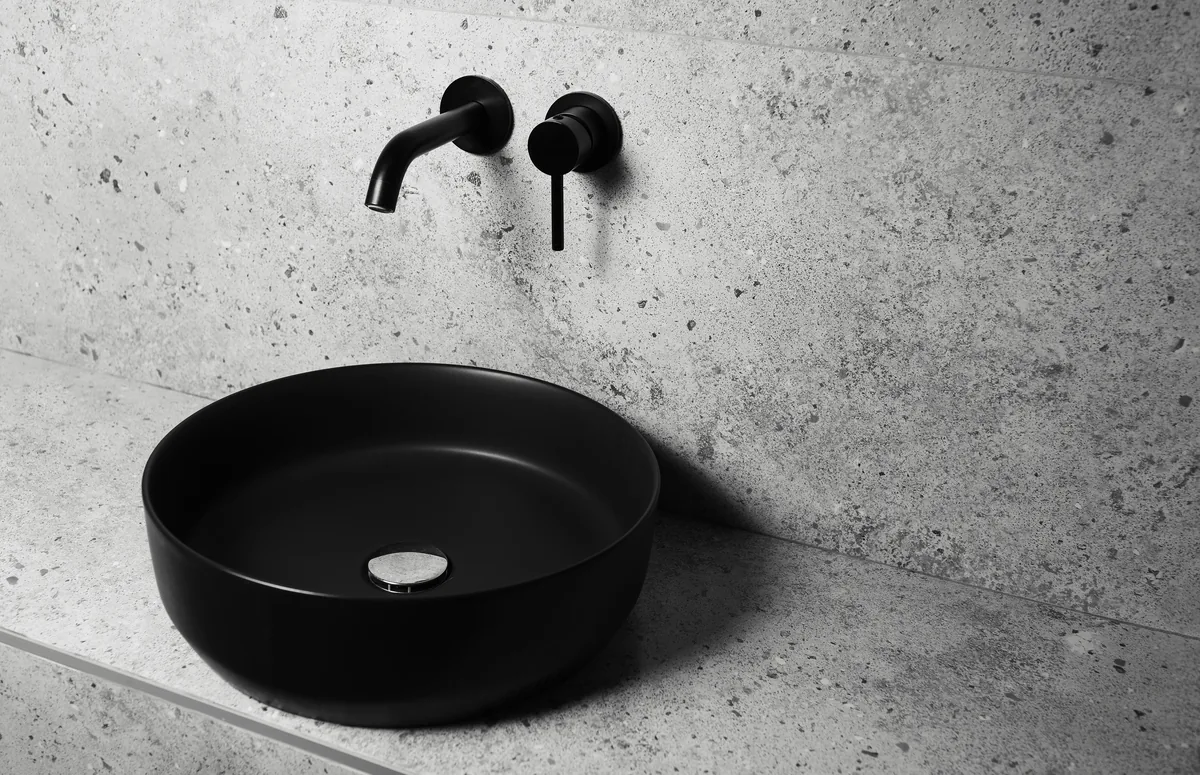 Trend arredo bagno 2027, superficie continua effetto cemento con lavabo tondo nero opaco e rubinetteria a parete in finitura opaca